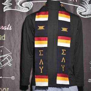May include: A black graduation stole with gold lettering that reads "ΣΔΥ" and a gold and white stripe pattern. The stole is worn over a black long-sleeved shirt.