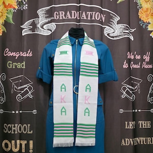 May include: White graduation stole with pink and green stripes and the letters "AKA" embroidered in pink and green. The stole is worn by a person wearing a blue dress. The background is a chalkboard with the words "Graduation", "Congrats Grad", "School Out!" and "We're off to Great Places" written on it.