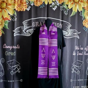 May include: A purple graduation stole with white stripes and Greek letters. The stole is displayed on a black background with the words "Congrats Grad" and "We're off to Great Places". The stole is a symbol of academic achievement.