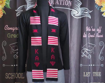 May include: A black graduation scarf with red and white stripes and the Greek letters K, A, and Ψ. The scarf is displayed on a black long-sleeve shirt and gray pants. The background includes text that says "Graduation" and "We're off to Great Places."