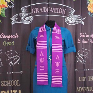 May include: Purple and pink graduation stole with the Greek letters Psi and Lambda. The stole has a white stripe pattern and the text "GRADUATION" is visible in the background.
