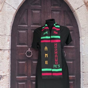 May include: Black scarf with green, red, and yellow accents. The scarf has the text "AME" and "MSHAWO PK'S" in yellow and green. The scarf is displayed on a black dress.