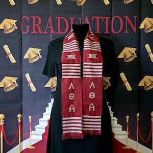 May include: A red and white graduation stole with the Greek letters "ΑΘΑ" in white. The stole has horizontal white stripes and decorative elements. The background features the word "GRADUATION" in red, along with graduation caps and scrolls.