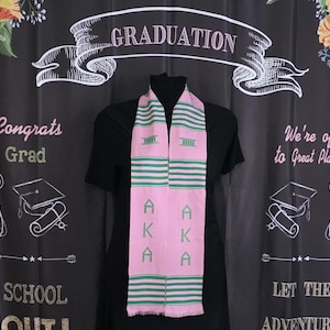 May include: A pink and green graduation stole with the letters "AKA" printed on it. The stole has green stripes and fringe at the bottom. The background includes the word "GRADUATION" and other graduation-themed text.
