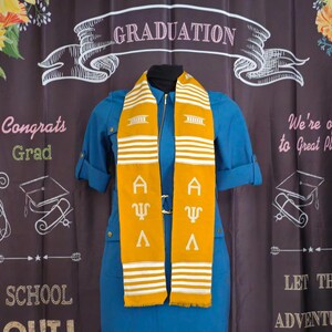 May include: A gold and white graduation stole with the Greek letters "A". "Ψ", and "Λ" embroidered on it. The stole is worn over a blue dress. The background is a black chalkboard with the words "GRADUATION", "Congrats Grad", "School Out", "We're off to Great Places", and "Let the Adventure Begin".