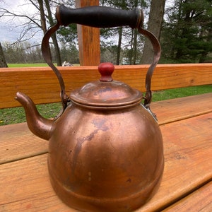 May include: A vintage copper tea kettle with a wooden handle and a red knob on the lid. The kettle is sitting on a wooden surface.