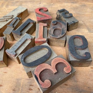 May include: A collection of vintage wooden letterpress blocks featuring various numbers and letters in different fonts and sizes. The blocks are made of light-colored wood with raised characters in black and red ink, showing signs of wear and age.