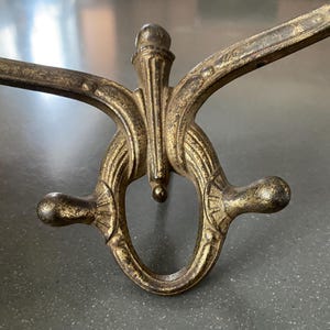 May include: Ornate, antique-style wall hook in a bronze finish. The hook features a decorative, symmetrical design with curved arms and a central, vertical element. The metal has a weathered appearance, suggesting age and use.