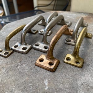 May include: A collection of six metal door handles arranged in two rows. The handles feature varying finishes, including polished brass, brushed nickel, and aged bronze tones. Each handle has a curved grip and two rectangular mounting plates.