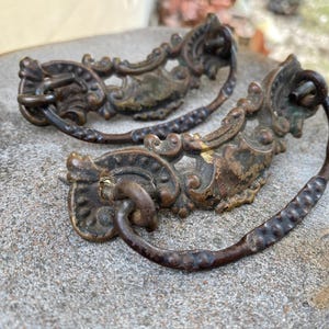 May include: Two antique bronze drawer pulls with ornate floral and scrollwork designs. The pulls have a dark, aged patina and feature a curved handle with a textured pattern. These vintage hardware pieces are likely from a cabinet or dresser.