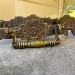 May include: A set of antique brass drawer pulls featuring a central six-pointed star design surrounded by intricate geometric patterns. Each rectangular pull has a hinged, rounded drop handle and decorative scalloped edges on both sides.