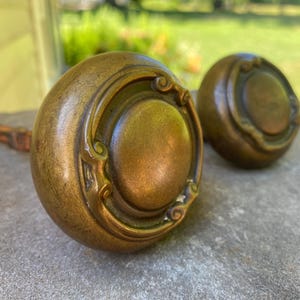 May include: Two round brass door knobs with a smooth central dome and a decorative raised border featuring curved scrollwork details. Each knob has a metallic finish with visible signs of age and patina, and one knob shows a square metal spindle.
