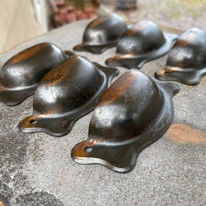May include: A collection of six antique metal drawer pulls. The pulls are a dark, aged bronze color with a rounded, domed shape and a small, curved tab with a hole for mounting. The pulls are arranged on a rough, gray surface.