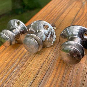 May include: Three silver-toned metal cabinet knobs with a rounded, mushroom-shaped head and a circular mounting base featuring two screw holes. The metallic finish shows signs of wear, including some white paint residue on the surfaces.