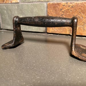 May include: A rustic, dark brown metal handle with a curved grip and two flat, foot-like bases. The handle appears aged with a textured surface, suggesting it is vintage or antique. The design is simple and functional.