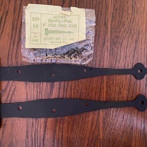 May include: Two black 9-inch strap hinges with decorative ends, along with a package of screws. The hinges are from Acorn Mfg. Co., and the package is labeled "Thrifty-Pak". The package also includes the text "9" Strap Hinge Flush".