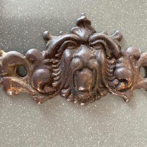 May include: A decorative, antique-style metal furniture pull with a lion's face design. The bronze-colored pull features intricate details and scrollwork, with two circular holes on either side. The piece has a weathered appearance.