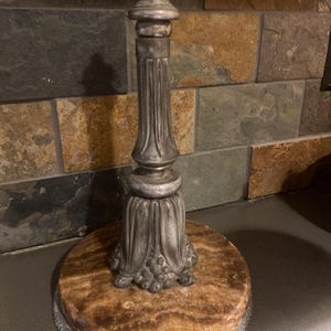 May include: A vintage lamp base with a round, marble-like base and a decorative, silver-colored metal stem. The base has a detailed, ornate design, and the stem features vertical grooves. The lamp base is designed to hold a lampshade.