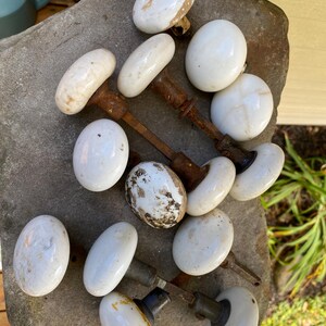 May include: A collection of vintage white porcelain door knobs with oval-shaped heads and weathered, rusted metal spindles. The knobs exhibit varying degrees of surface wear, including brown staining, cracks, and dark discoloration on the ceramic.