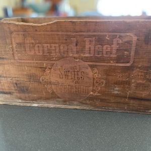 May include: A vintage wooden crate with the words "Corned Beef" and "Swift's Premium" embossed on the front. The crate is made of weathered wood, showing signs of age and use. The crate is rectangular and has a rustic appearance.