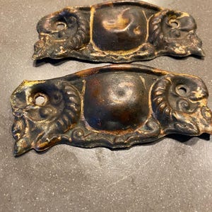 May include: Two antique brass drawer pulls with ornate designs. The pulls are shaped like a rounded rectangle with a raised center and intricate details on the edges.