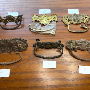 May include: Six antique cabinet hardware pulls with ornate designs. The pulls are made of brass and have a variety of finishes, including polished brass, aged brass, and black.