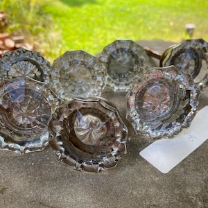 May include: A collection of six clear glass door knobs featuring a faceted, starburst-like pattern on the front surface. Each knob has a circular, scalloped edge design and a metallic base, arranged together on a flat, textured stone surface.
