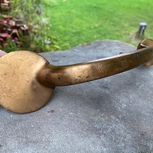 May include: A curved brass drawer pull featuring a smooth, arched handle connected to two circular mounting plates. The metal surface displays a weathered, aged patina with subtle variations in color and texture across the entire hardware piece.