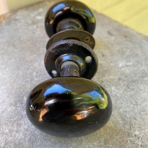 May include: A pair of round, glossy black door knobs featuring a smooth, reflective surface. The knobs are connected by a central metal spindle and include two circular metal mounting plates with visible screw holes, showing signs of age and wear.