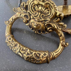 May include: Close-up of antique brass drawer pulls with ornate, floral designs. The pulls have a ring handle and decorative backplates. The brass has a warm, aged patina, suggesting vintage or antique hardware. The hardware is likely for cabinets or drawers.