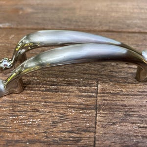 May include: Two silver-colored cabinet handles with a curved design. Each handle has a decorative end piece and a smooth, arched grip. The handles are displayed on a wooden surface.