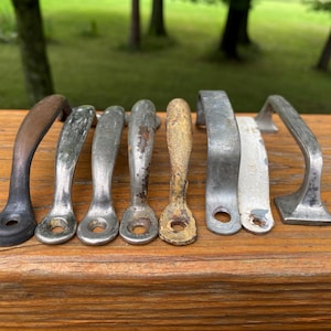 May include: A collection of eight metal drawer pulls and cabinet handles in various finishes, including aged bronze, weathered silver, tarnished gold, and white paint. Each handle features a curved design with circular mounting holes at the ends.