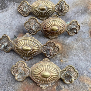 May include: Four antique brass drawer pulls. Each pull features an oval center with a sunburst design, flanked by ornate floral-shaped ends. The pulls are aged with a warm, golden hue, suggesting vintage craftsmanship. Ideal for furniture restoration or adding a vintage touch.