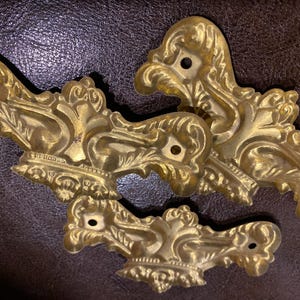 May include: Four ornate, gold-colored furniture hardware pieces with intricate scrollwork and a small hole for mounting. The hardware has a vintage aesthetic and is likely made of brass or a similar metal. The pieces are arranged on a dark brown surface.