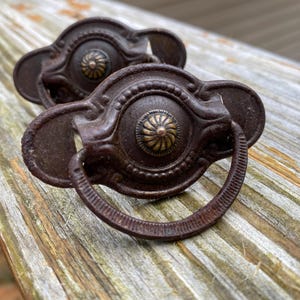May include: Two antique bronze drawer pulls with a decorative design. Each pull features a central medallion with a floral pattern and a ring handle. The pulls are aged with a rustic finish, perfect for vintage furniture restoration.