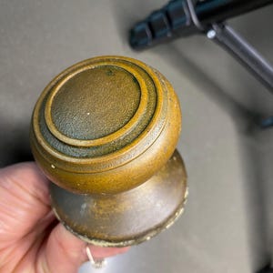 May include: Antique brass doorknob with a round, ridged design. The doorknob has a weathered, aged appearance, with visible signs of wear and patina. The hardware is held in a hand, suggesting its size and use.