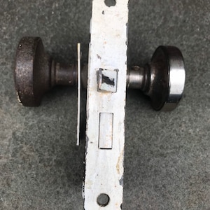 May include: An antique door lock set with a white, rectangular faceplate and two dark brown, round doorknobs. The faceplate shows signs of wear with chipped paint. The doorknobs are made of metal and show signs of age.