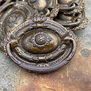 May include: Close-up of antique bronze drawer pulls with ornate detailing. The pulls feature a central oval design with a floral motif and a curved handle. The bronze finish has a distressed look, adding to the vintage aesthetic.