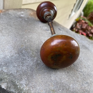 May include: A pair of round, dark brown, polished door knobs connected by a central metal spindle. The knobs feature a smooth, spherical shape with visible signs of wear and aging on the surface, showcasing a deep, warm, and glossy brown finish.
