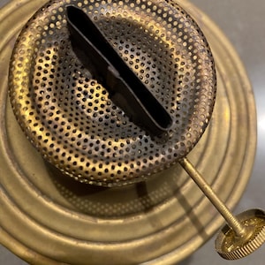 May include: Brass lamp burner with a perforated metal cap and a black metal wick holder. The burner is attached to a brass base with a threaded knob.