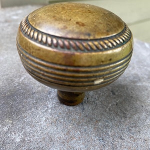 May include: A round, brass-toned metal cabinet knob featuring a weathered, aged patina. The surface displays a decorative rope-like texture around the upper edge, followed by three horizontal grooved lines circling the main body of the knob.