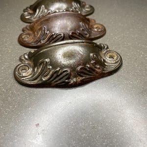 May include: Three antique drawer pulls with ornate, detailed designs. The pulls are made of metal and have a dark, aged finish.