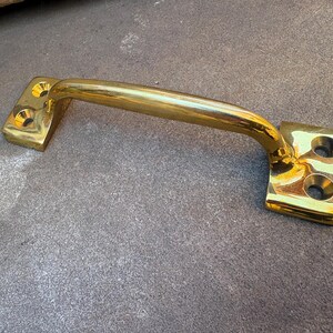 May include: A polished brass door handle featuring a curved grip and two rectangular mounting plates at each end. Each mounting plate contains two circular screw holes for installation, providing a secure and functional hardware component.