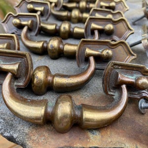 May include: A collection of antique brass drawer pulls. Each pull features a curved handle and a decorative backplate with a stylized design. The brass has a warm, aged patina, with some showing signs of wear. The pulls are likely for furniture restoration.