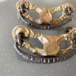 May include: Two antique bronze drawer pulls with ornate floral detailing and a dark brown handle. The pulls have a curved shape and are designed to be mounted on a drawer or cabinet. The hardware has a vintage aesthetic.
