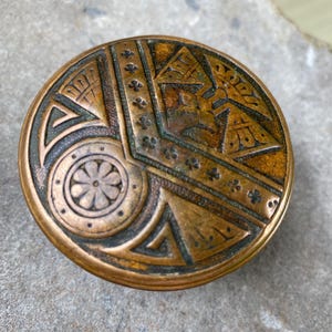 May include: A round, metallic bronze-toned object featuring an intricate, engraved geometric pattern. The design includes a central circular floral motif, triangular shapes, and a linear border decorated with small, repeating cross-like symbols.