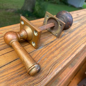 May include: A vintage door hardware set featuring a brass lever handle attached to a square mounting plate, connected by a metal spindle to a round, dark brown wooden door knob with a matching square mounting plate and visible screw holes.