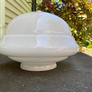 May include: A glossy white glass light fixture shade with a smooth, rounded dome top and a stepped, tiered profile that tapers down to a circular base opening. The surface is reflective, showing light highlights across its curved, opaque structure.