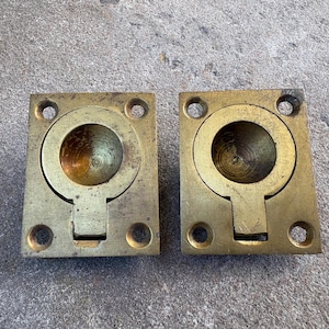 May include: Two rectangular brass flush mount ring pulls featuring a central circular recessed area with a hinged drop-down handle. Each piece has four pre-drilled screw holes at the corners, showing a weathered metallic finish and patina.