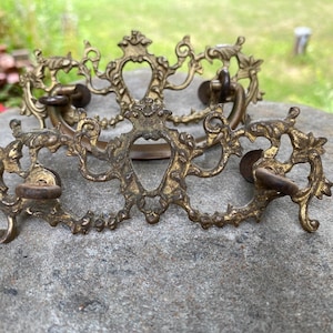 May include: Two ornate brass drawer pulls featuring intricate scrollwork and leaf motifs. Each piece has a central decorative plate with a curved bail handle attached by two circular mounting points, showcasing a weathered, aged metallic finish.